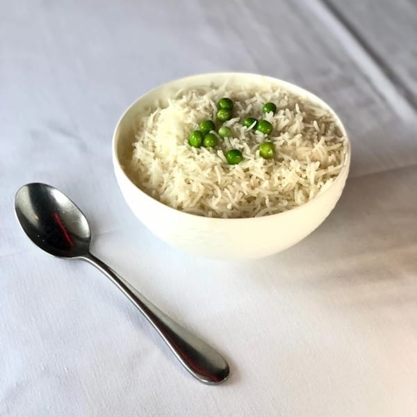 A white bowl of basmati rice with green peas, next to a silver spoon on a white tablecloth.