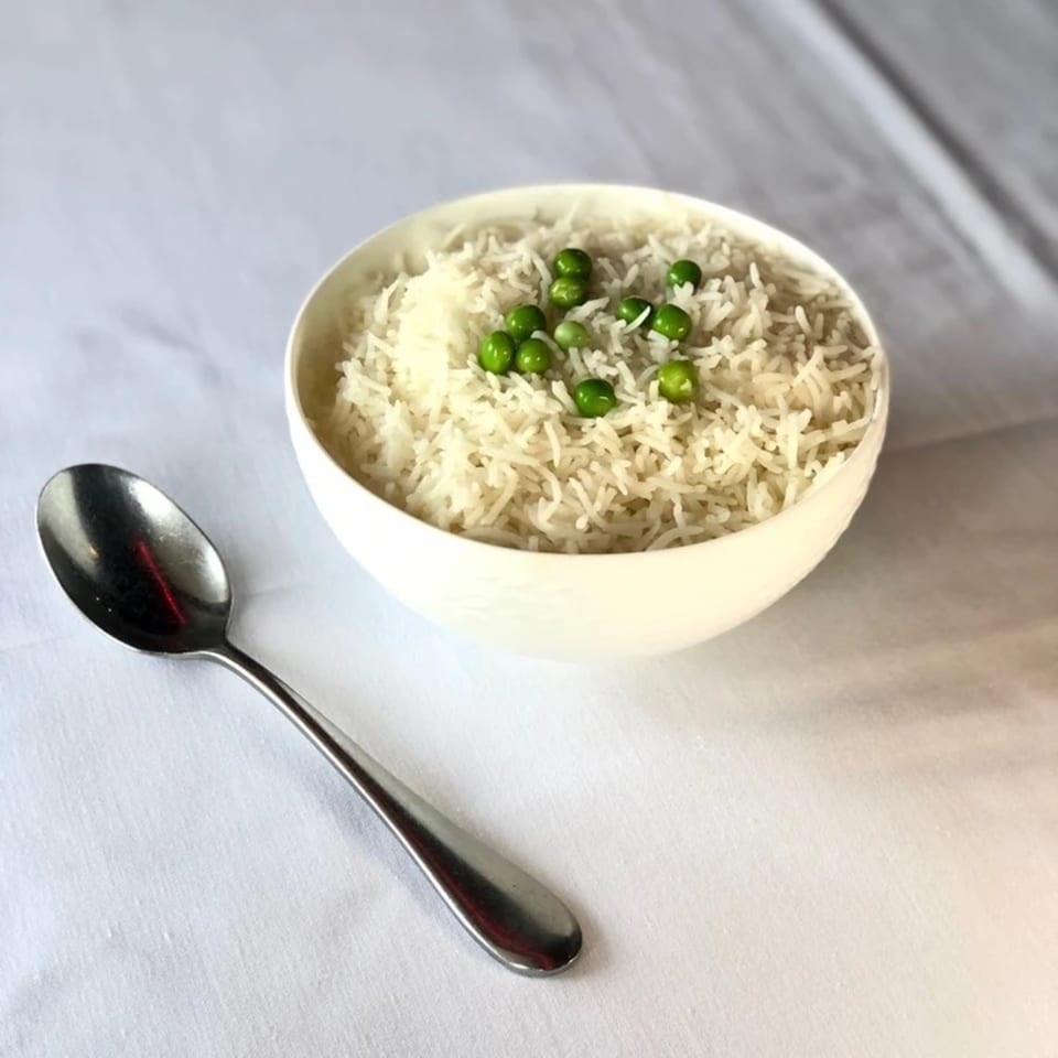 A white bowl of basmati rice with green peas, next to a silver spoon on a white tablecloth.